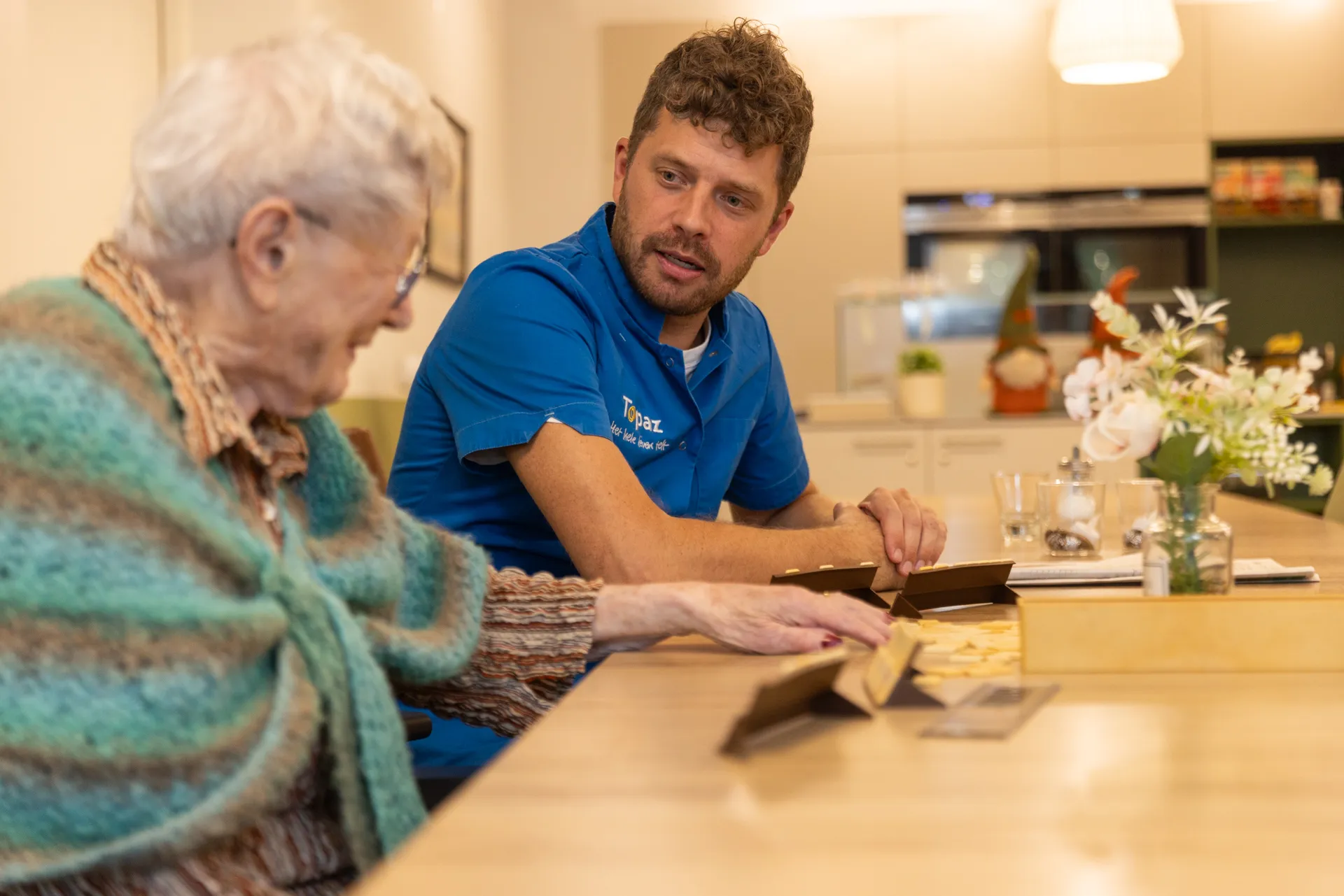 Zorgcollega van Topaz zit met een bewoner aan tafel Zorgcollega van Topaz zit met een bewoner aan tafel