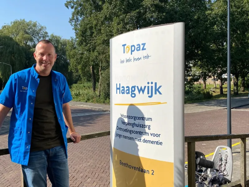 Zorgmedewerker die buiten naast het bord van Topaz Haagwijk staat.