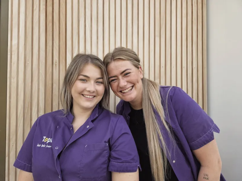 Twee zorgmedewerkers in paarse uniformen die lachend naast elkaar staan.
