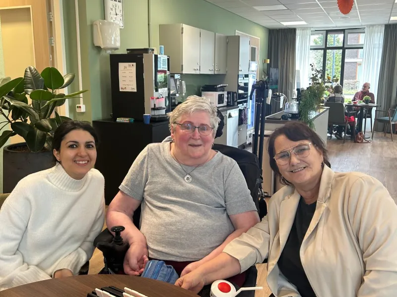 Bewoner in een rolstoel die samen met twee bezoekers aan een tafel zit en glimlacht.