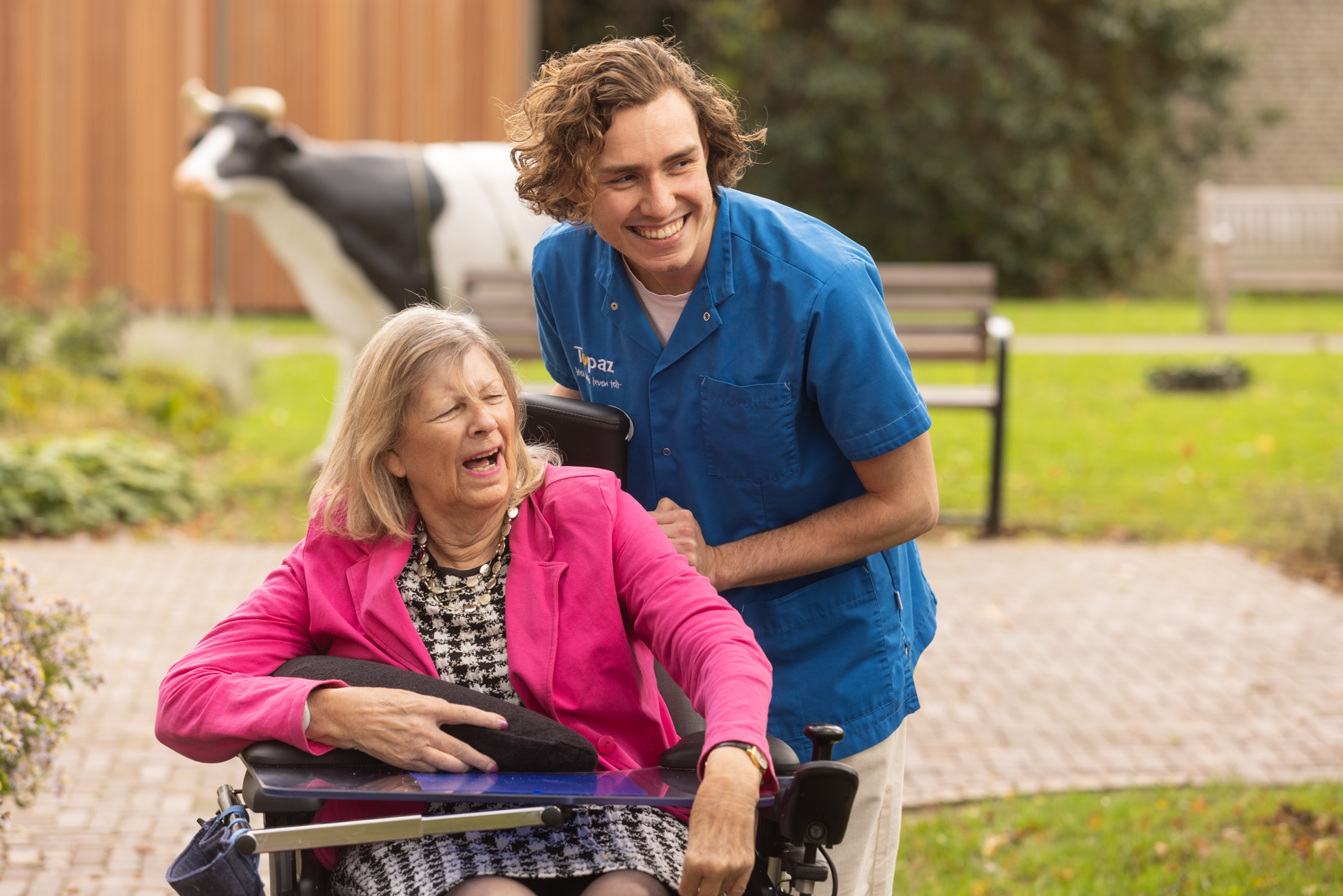 Zorgmedewerker die buiten met een lach een vrouw in een rolstoel begeleidt.