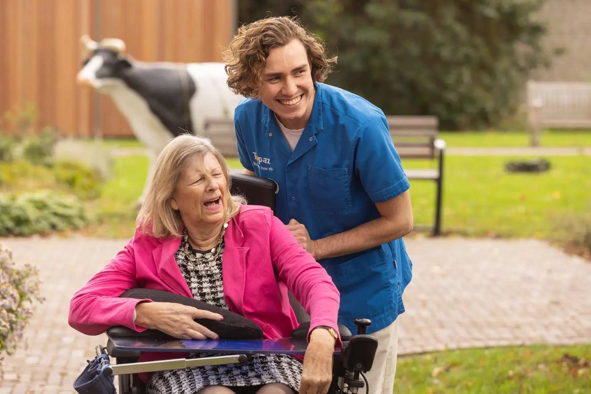 Zorgmedewerker die buiten met een lach een vrouw in een rolstoel begeleidt.